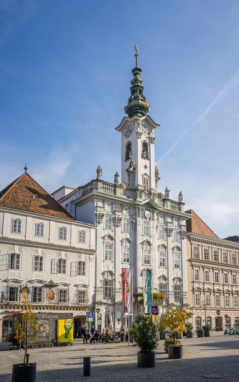 Historisches Gebäude mit Uhrturm auf einem sonnigen Platz, umgeben von weiteren Gebäuden und Pflanzen. Einladendes Stadtbild mit architektonischen Details und Flaggen im Vordergrund.