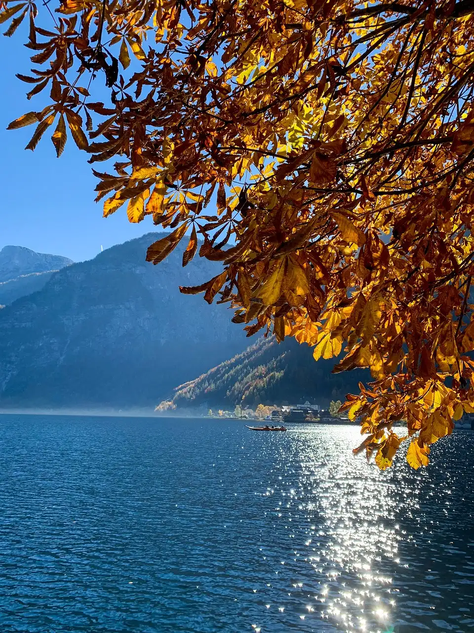 Herbstlandschaft mit einem See, umgeben von Bergen im Hintergrund, leuchtenden orange-gelben Baumblättern im Vordergrund und einem kleinen Boot auf dem glitzernden Wasser.