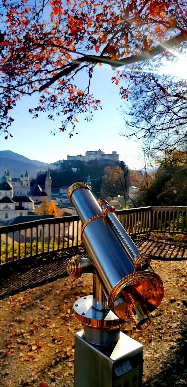 Silbernes Teleskop auf einem Aussichtspunkt mit Blick auf eine historische Stadt, Burg auf einem Hügel und herbstliches Baumlaub.